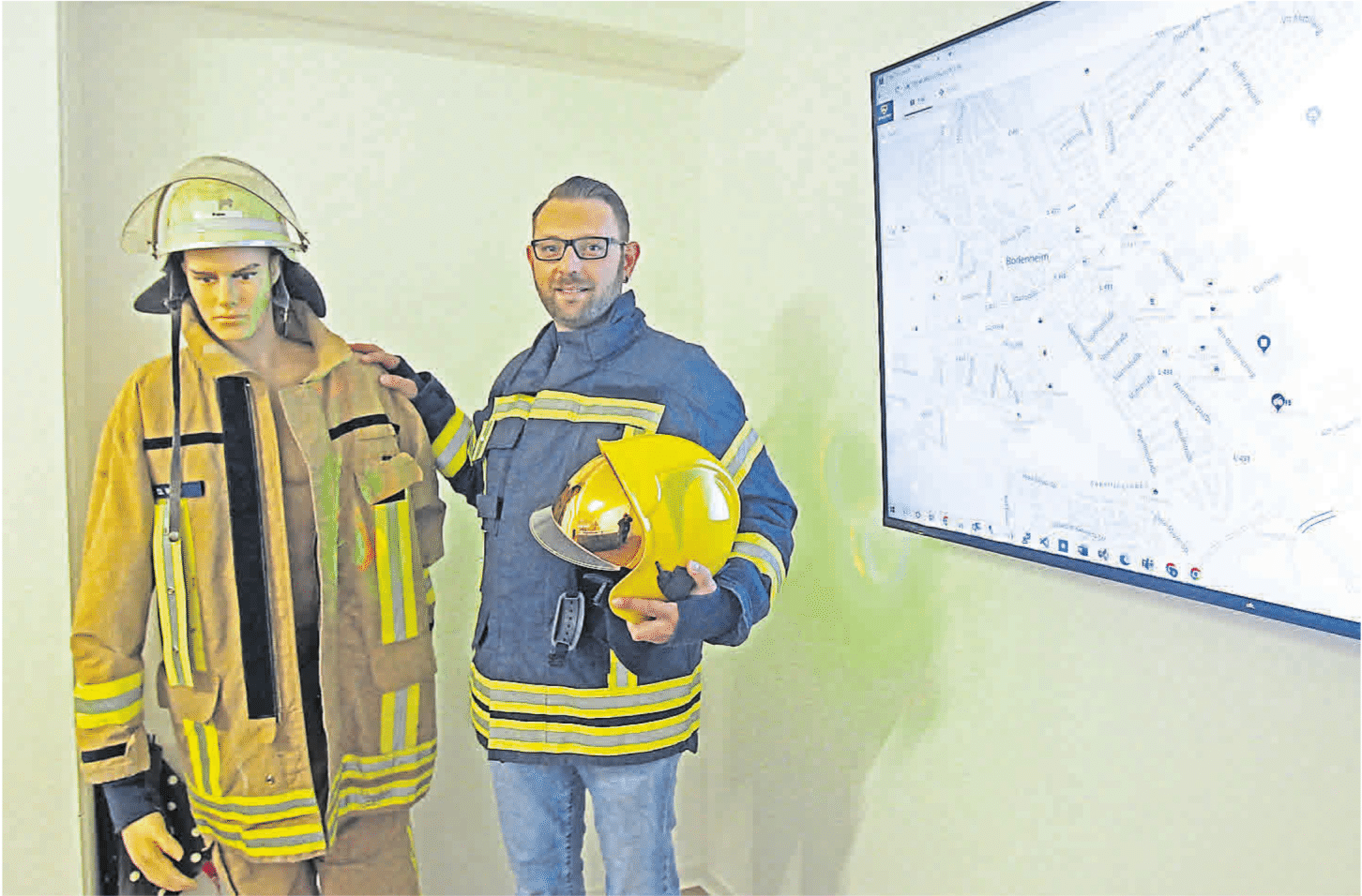 Denis Weber zeigt eine Feuerwehrjacke neben einer Schaufensterpuppe mit Schutzkleidung und einem Bildschirm zur Standortdarstellung.